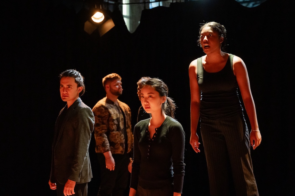 Four actors stand on stage with faces lit from below in mid-performance of The Winter's Tale. The actor on the right is in mid-speech as the others look forward sternly.